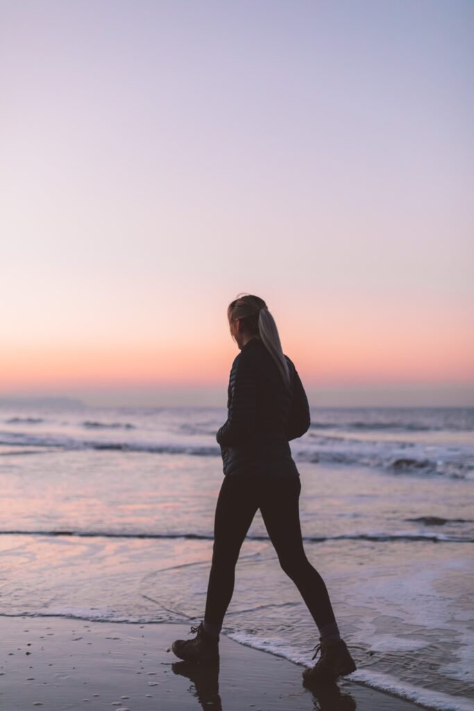 Woman walking alone along a calm shoreline, facing forward.
