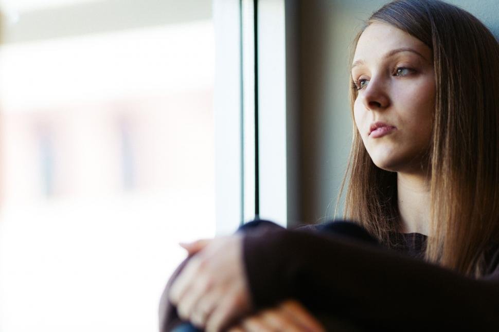 Woman sitting by a window in soft light, looking thoughtful and reflective.