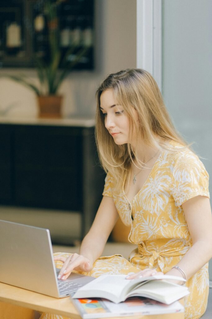 Young woman in a yellow dress working on a laptop with a book open indoors.