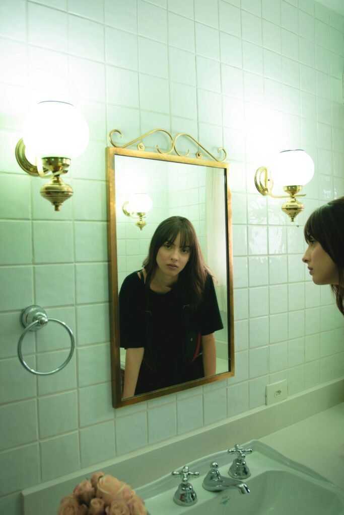 Woman looking into a bathroom mirror next to vintage lamps.