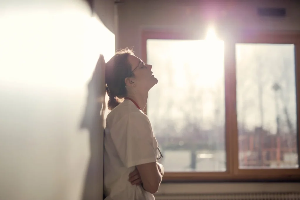 Woman standing quietly leaning against the wall in soft natural light, reflecting emotional exhaustion and deep thought.